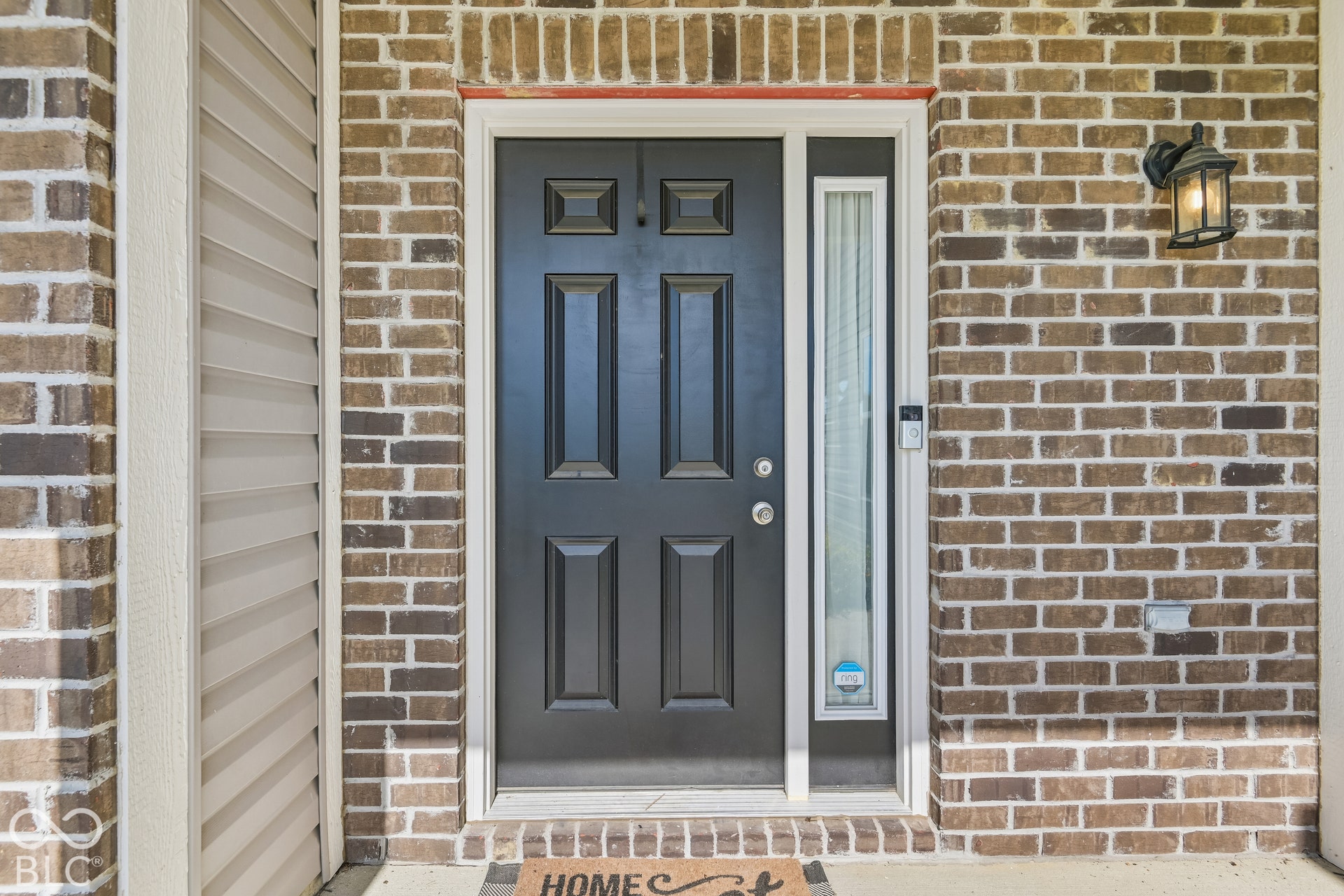Front entryway featuring a dark six-panel entry door, vertical sidelight window, multi-tonal brick facade, and exterior wall-mounted lantern lighting and a Ring Doorbell