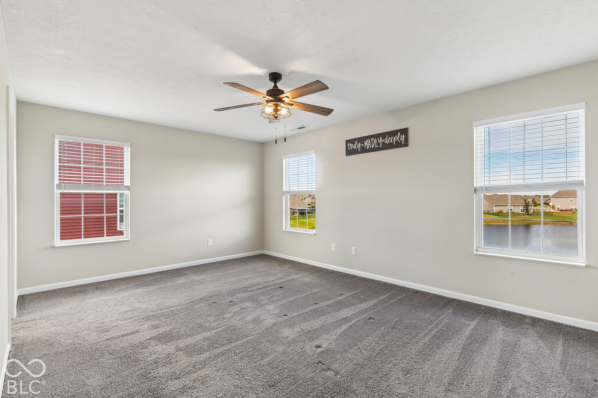Neutral wall tones, wall-to-wall carpeting, three windows with blinds, and a multi-blade ceiling fan with light fixture
