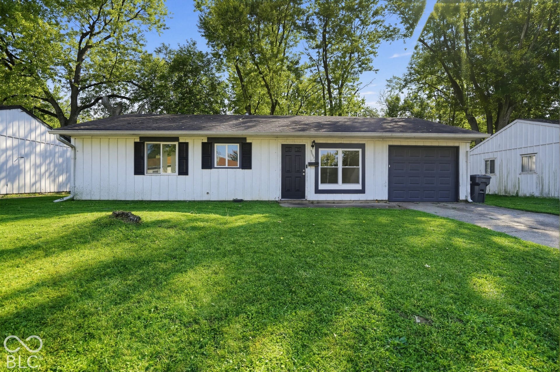 Single-story residence featuring vertical siding, dark-trimmed windows, a front-entry door with glass detail, an attached single-car garage, and mature trees on the lot