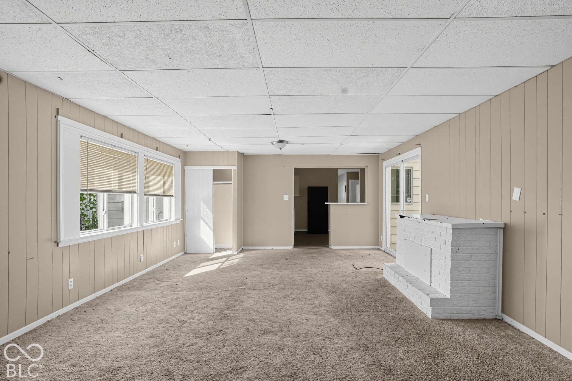Spacious interior featuring a white painted brick hearth, large windows with installed blinds, sliding glass doors, and a drop ceiling