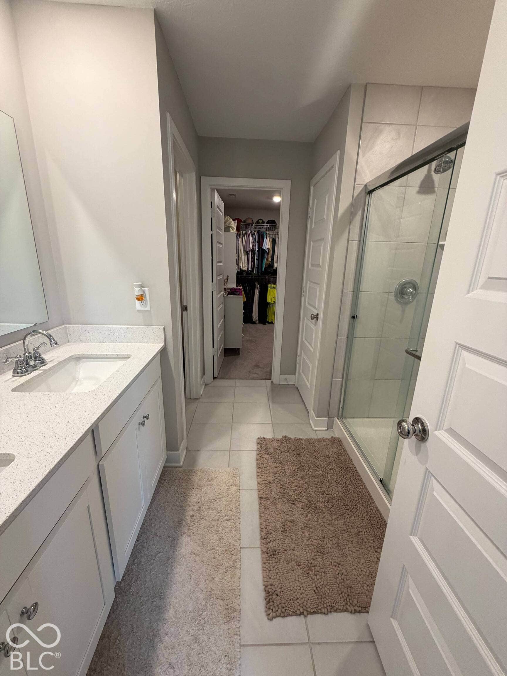 Vanity with white cabinetry and light-toned countertop, sink with chrome faucet, glass-enclosed shower with tiled walls, and tiled flooring