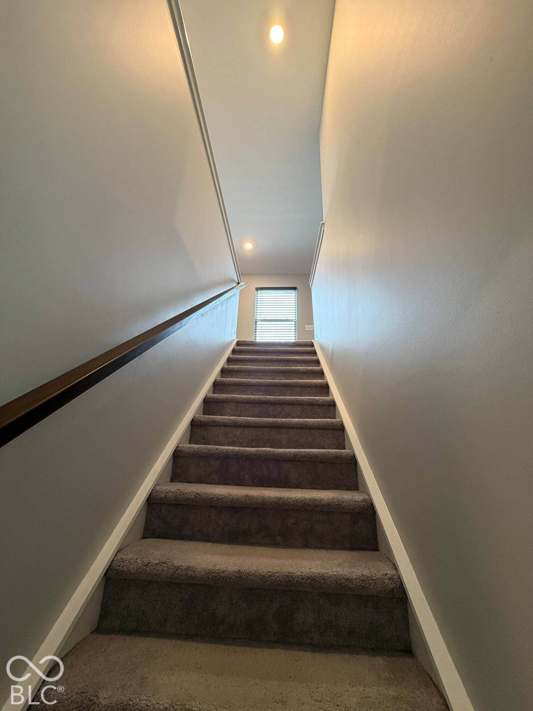 Carpeted staircase with a wood handrail, recessed lighting, and a window with blinds
