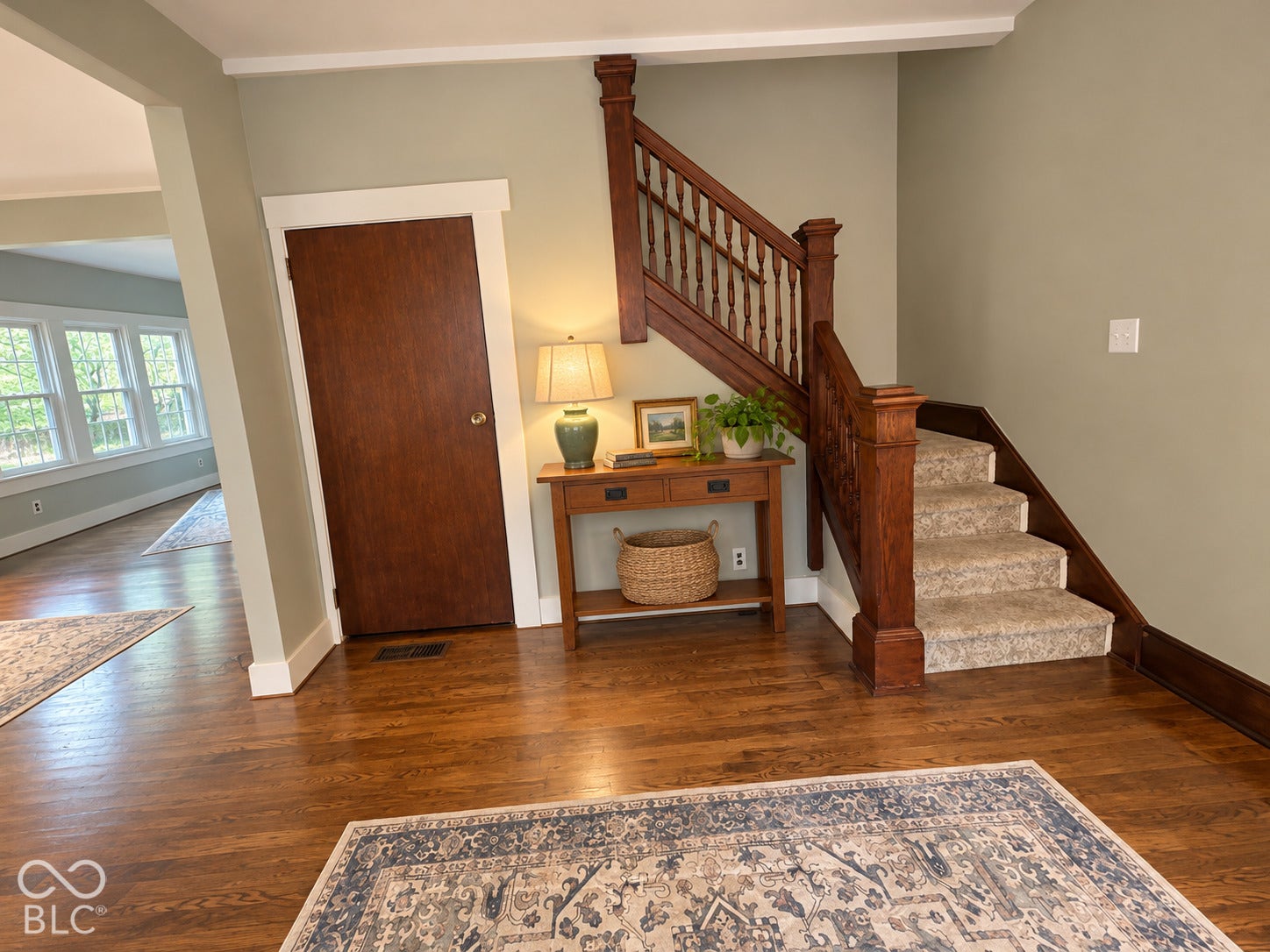 Virtually Altered - Entryway featuring wood-finish flooring, a solid wood door with decorative trim, and a wooden staircase with carved balusters and carpeted treads