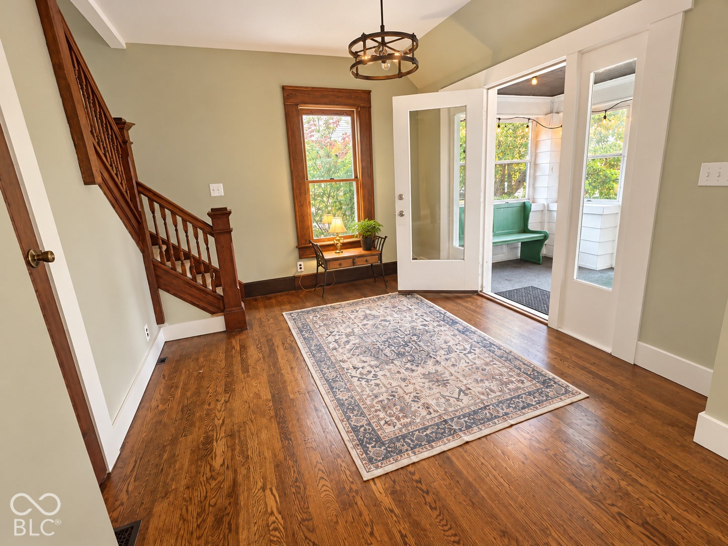 Wood-finish flooring, natural wood staircase with decorative balusters, craftsman-style window casing, and glass-paneled door opening to a covered porch with pendant lighting