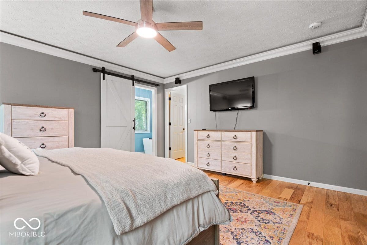 Primary bedroom featuring wood-finish flooring, crown molding, a ceiling fan with integrated lighting, and ensuite bathroom and walk-in closet