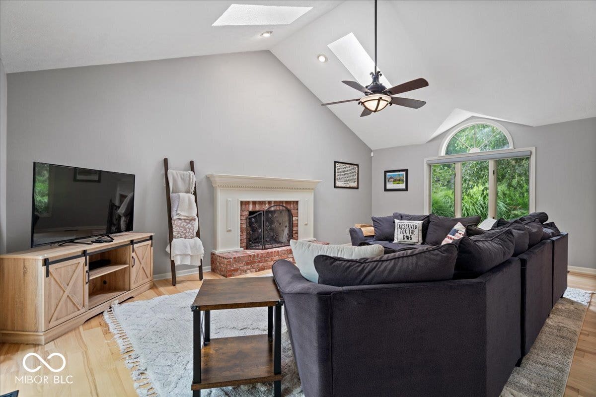 Vaulted ceilings featuring dual skylights, red brick fireplace with a decorative mantel, wood flooring, and an arched window
