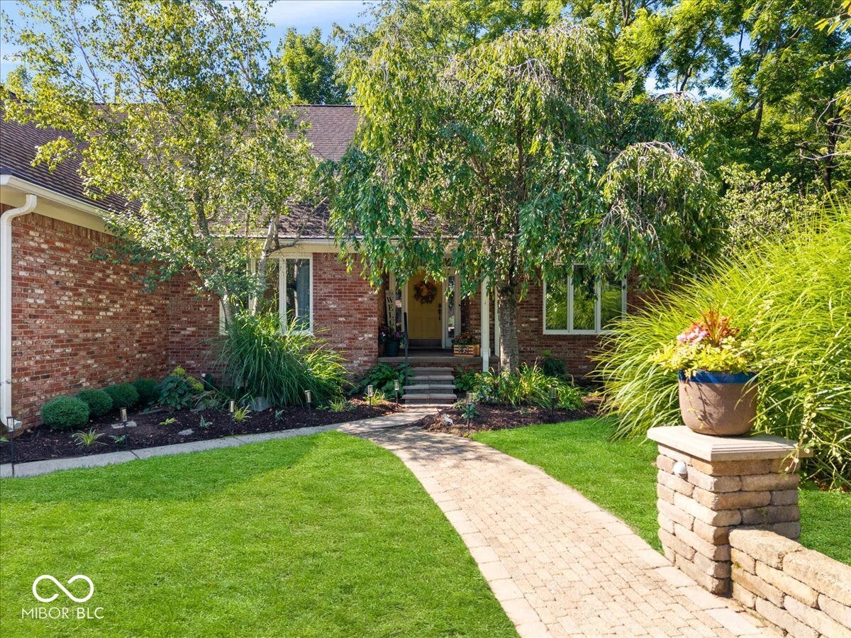 Brick exterior facade with a stone-paved walkway, tiered landscape beds, mature trees, and a low-profile stacked stone wall