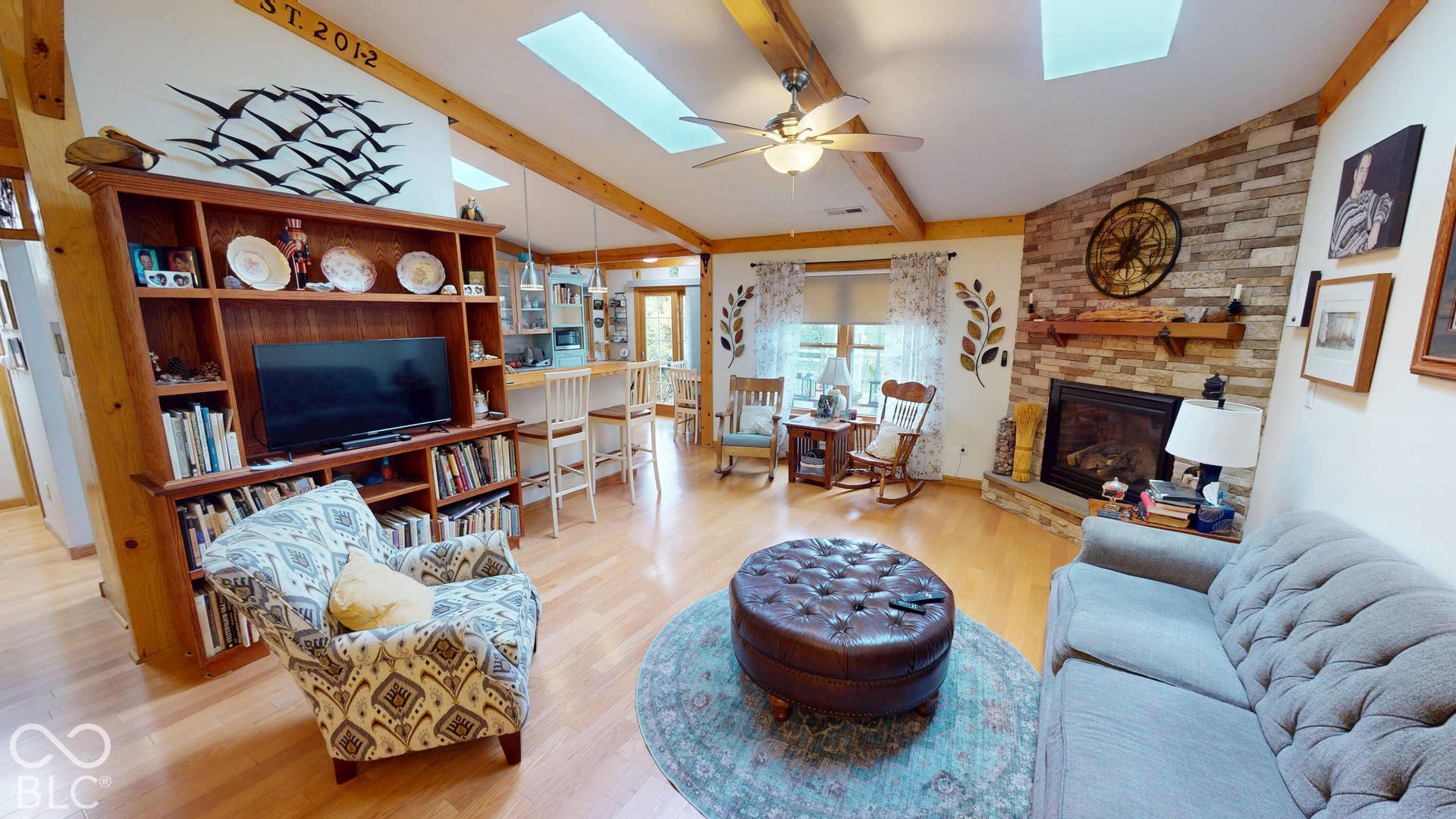 Vaulted ceiling with exposed wood beams, wood-finish flooring, stone-faced fireplace, built-in shelving, and skylights
