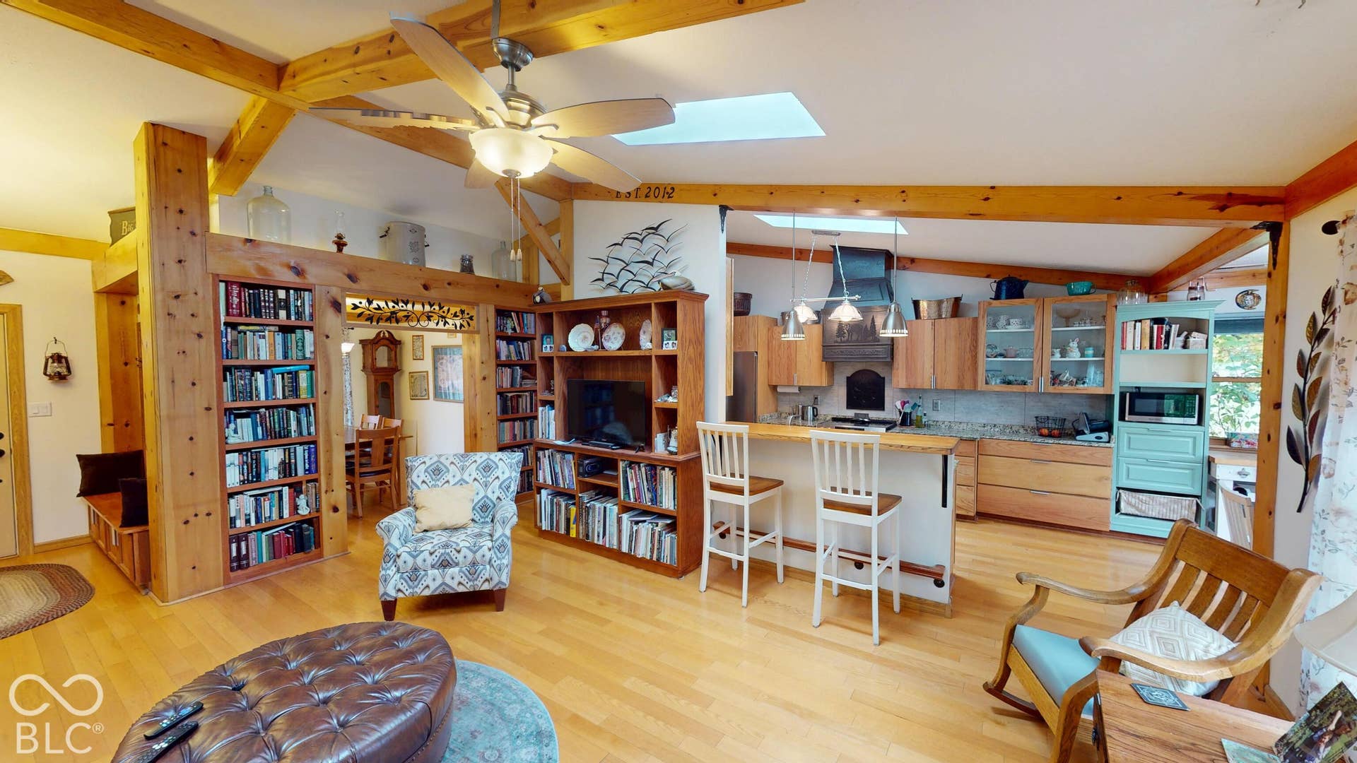Vaulted ceiling with exposed wood beams, wood-finish flooring, built-in shelving, kitchen with light wood cabinetry, and a skylight