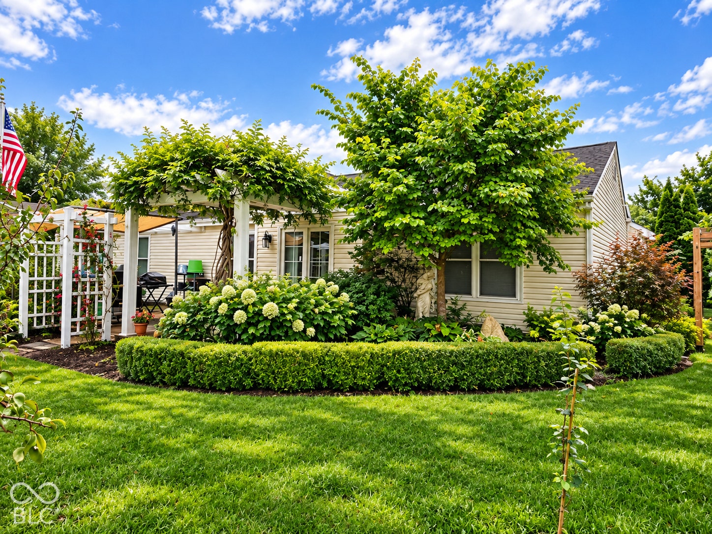 Expansive lawn with mature landscaping, featuring a white pergola with climbing greenery, defined garden beds, and a perimeter of manicured hedges