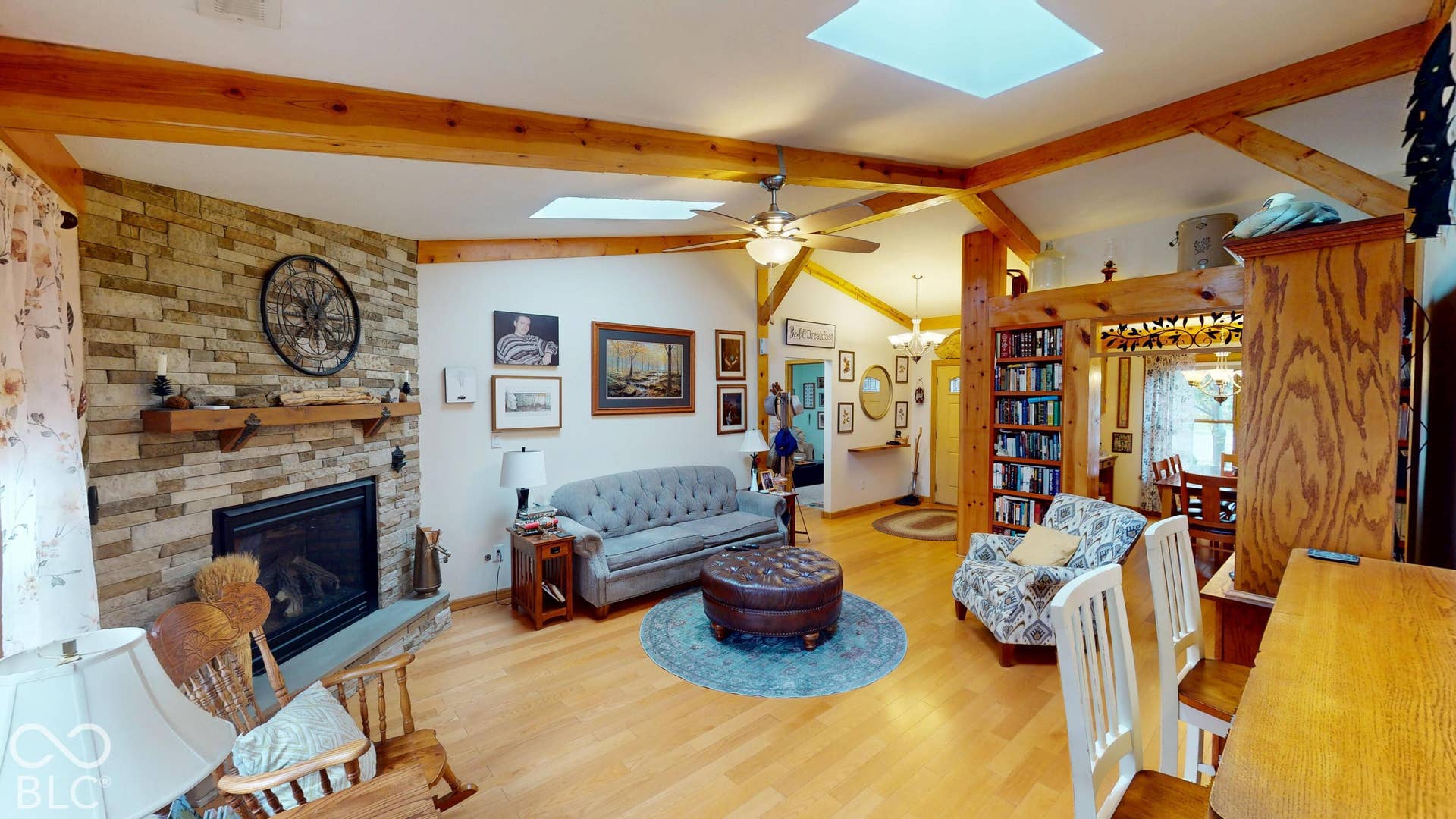 Living space featuring a stone-clad fireplace with a wood mantel, wood-finish flooring, exposed wood beams, and a ceiling fan
