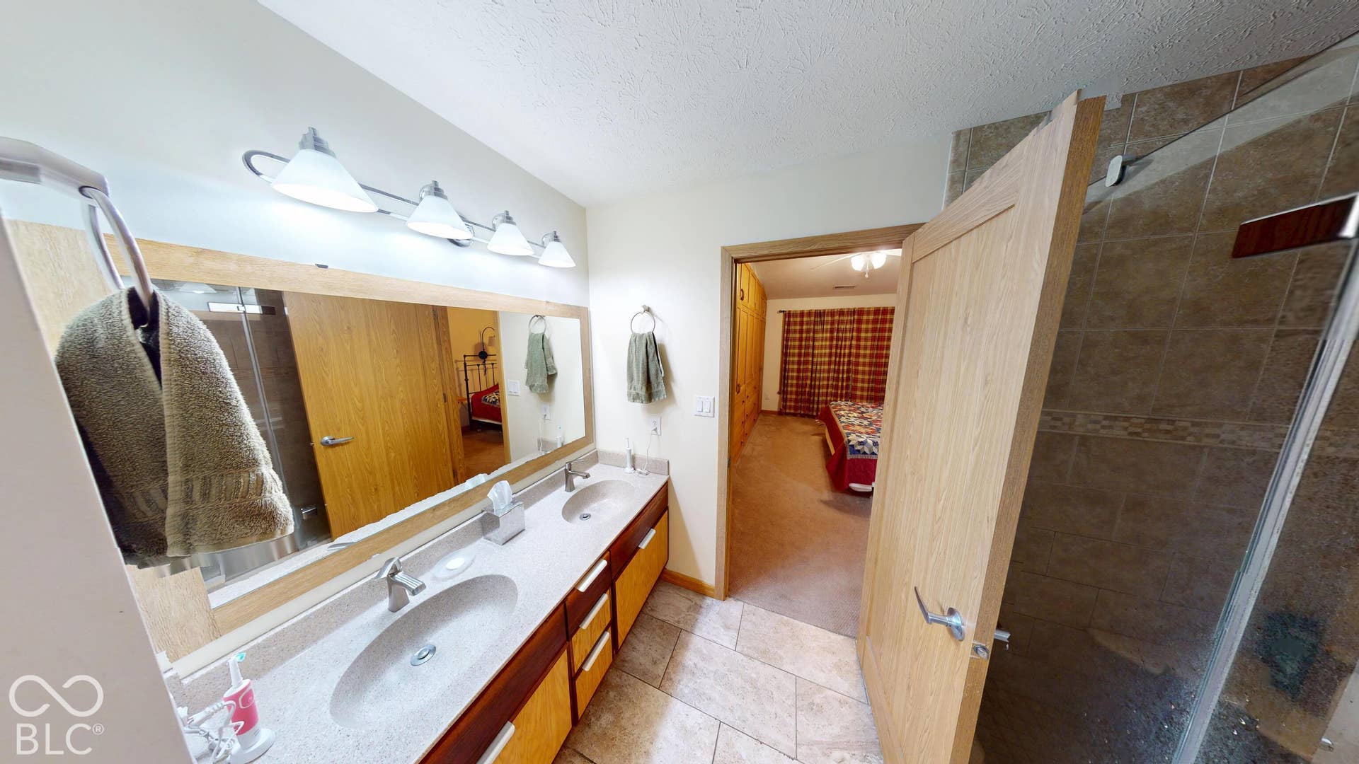 Dual vanity with integrated sinks, tiled flooring, wood-finish cabinetry, a large vanity mirror, and a tiled shower enclosure