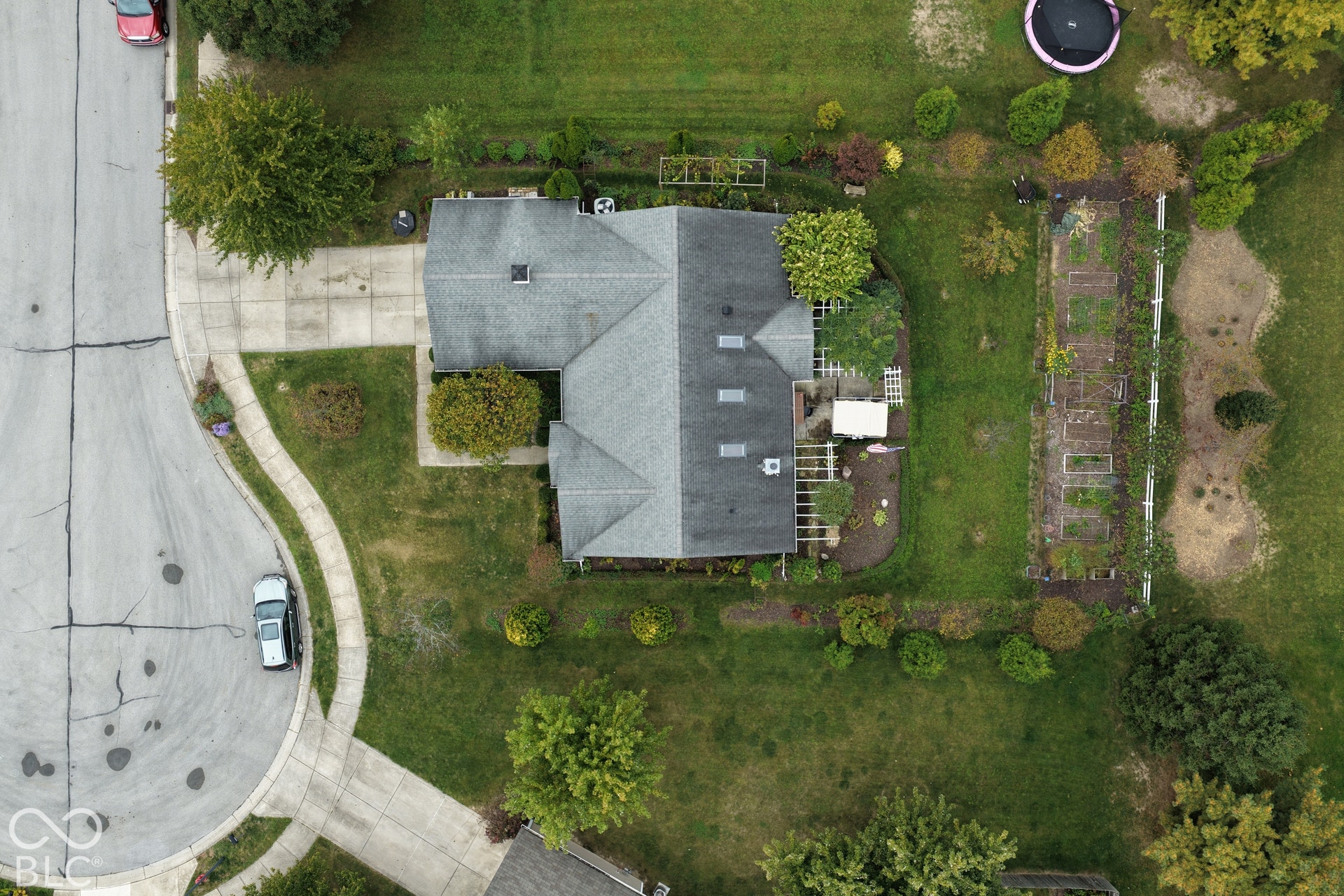Aerial view of a property featuring a multi-pitched roofline with skylights, a paved driveway, and an expansive lot with established garden beds and a wooden pergola