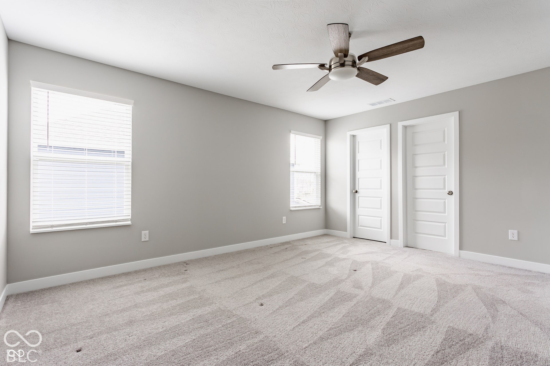 Neutral-toned interior space featuring wall-to-wall carpeting, two multi-panel doors, dual windows with fitted blinds, and a ceiling-mounted fan with wood-finish blades