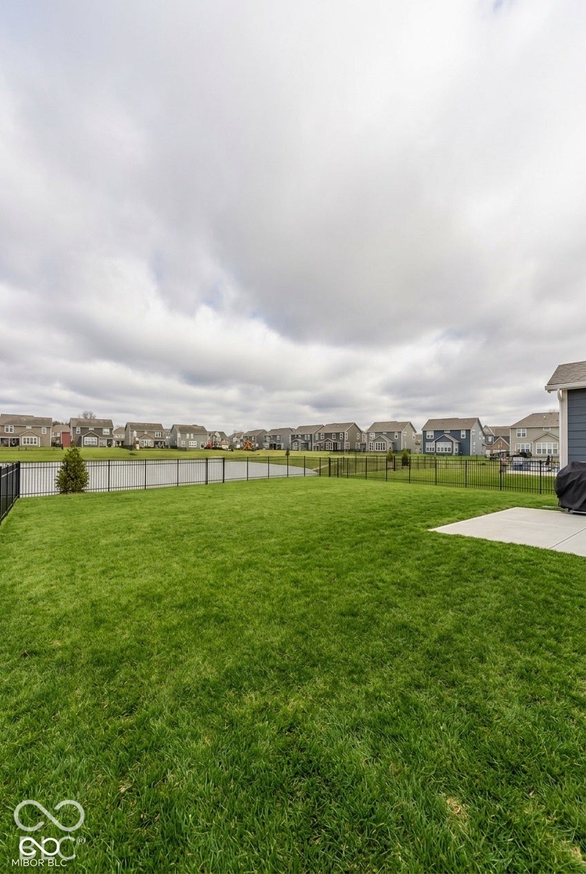 Rear yard featuring a manicured lawn, concrete patio, and black metal perimeter fencing, bordered by a pond