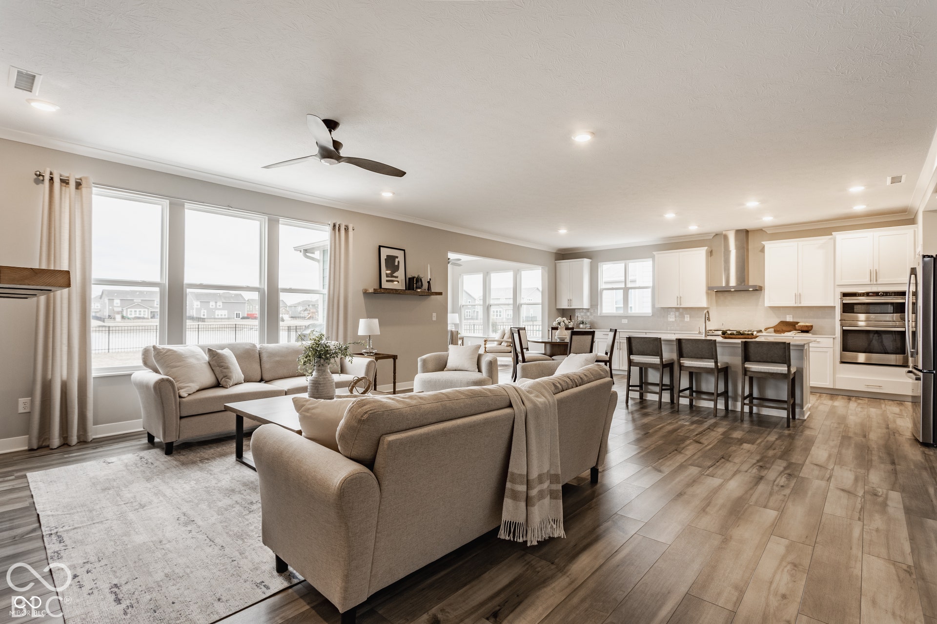 Open-concept interior featuring wood-finish flooring, white shaker-style cabinetry, stainless steel built-in double wall ovens, recessed lighting, and large windows