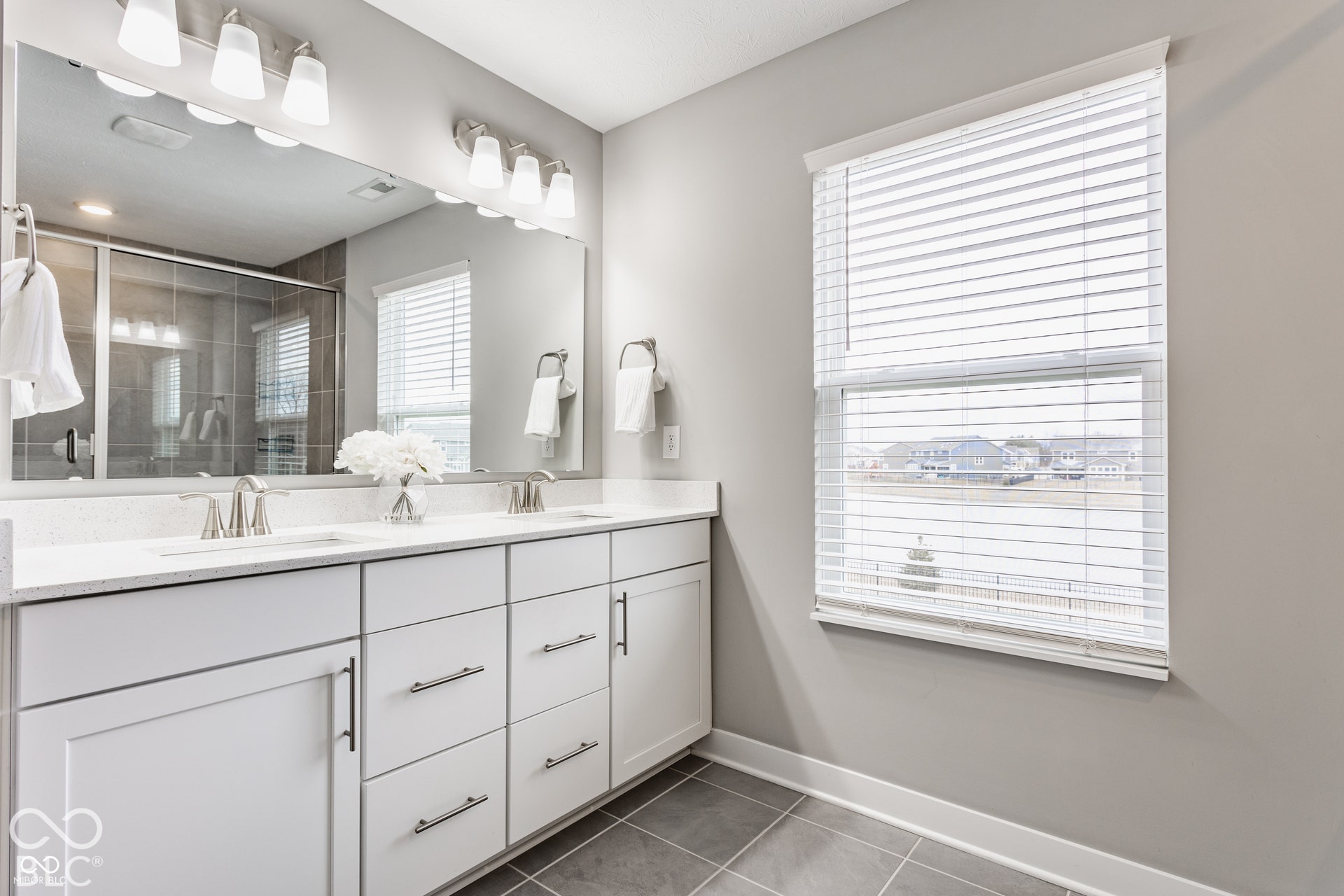 Dual-sink vanity with white cabinetry, light-toned stone-finish countertop, polished chrome fixtures, full-width wall mirror, and large-format tile flooring