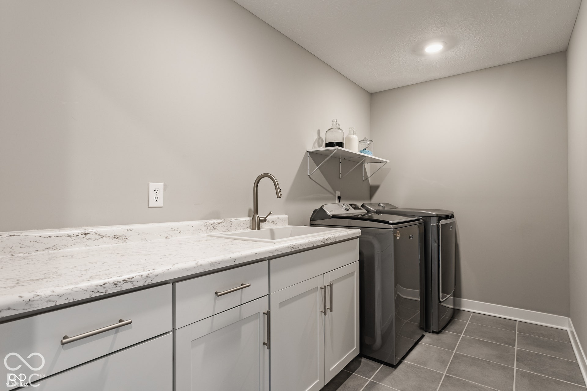 Laundry room featuring light grey cabinetry, stone-patterned laminate countertop, integrated drop-in sink with high-arc faucet, and tiled flooring