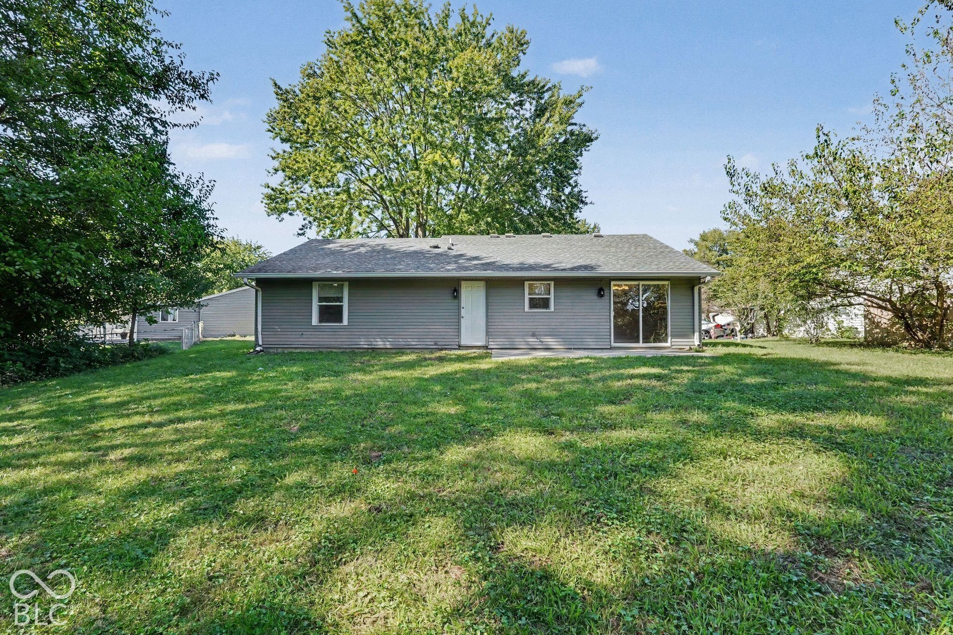 Expansive grassy yard with mature trees, gray siding exterior, white entry door, and a sliding glass door
