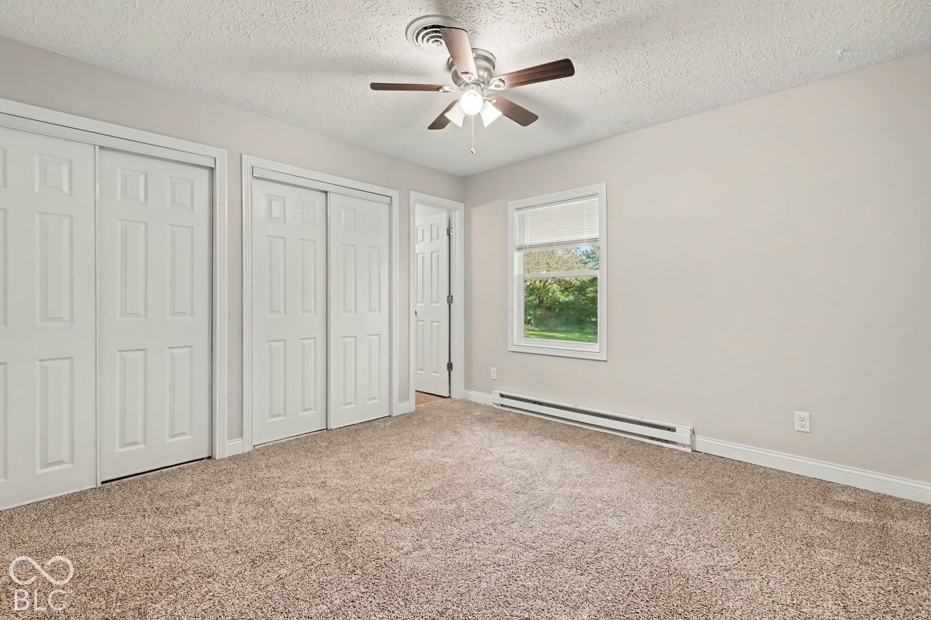 Neutral-toned carpeting, ceiling fan with light fixture, window with roller shade, and two sets of bi-fold closet doors