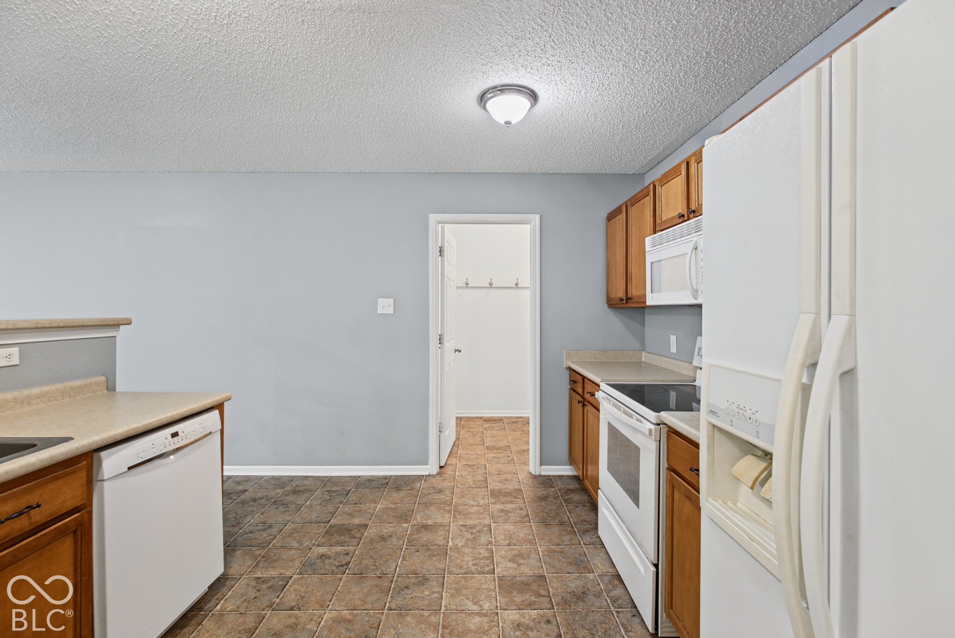 Kitchen featuring medium-tone wood cabinetry, solid-surface countertops, tile flooring, and white built-in appliances including a refrigerator, dishwasher, range, and over-the-range microwave