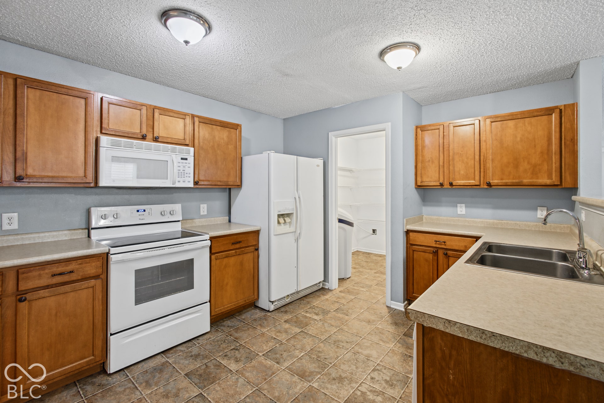 Kitchen featuring wooden cabinetry, white appliances, a double-basin stainless steel sink, tiled flooring, and a walk-in storage area