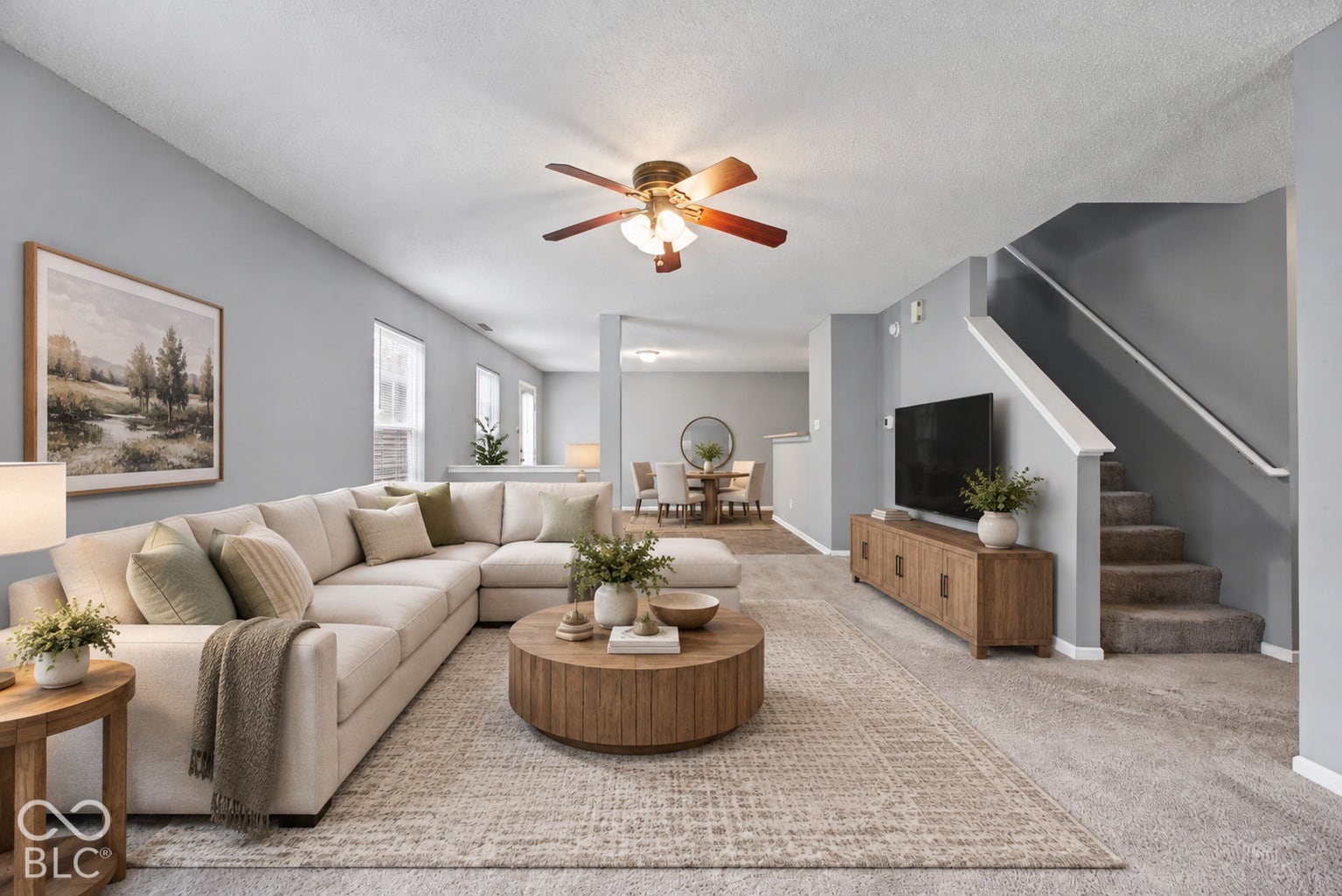 Virtually Altered - Neutral-toned living space featuring wall-to-wall carpeting, vaulted ceilings, a central ceiling fan with integrated lighting, and a staircase with a painted handrail