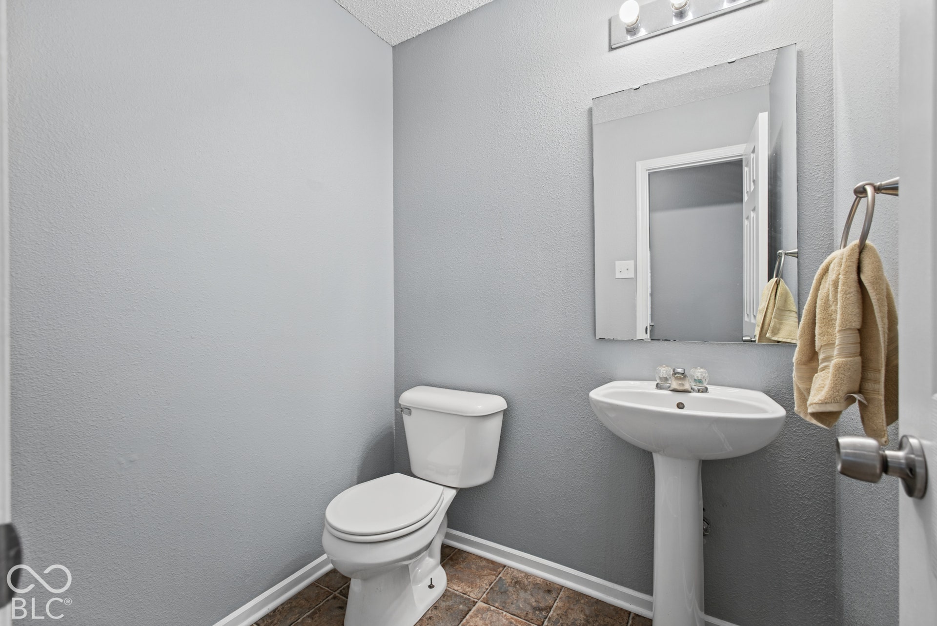 White pedestal sink with a dual-handle faucet, wall-mounted mirror, three-light fixture, and ceramic tile flooring