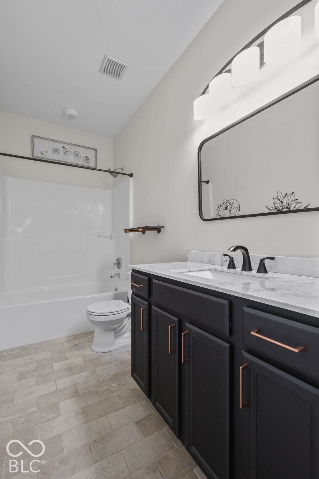 Dark-toned vanity with metallic hardware, light-toned stone-finish countertop, undermount sink with widespread faucet, rounded-corner wall mirror, and porcelain tile flooring