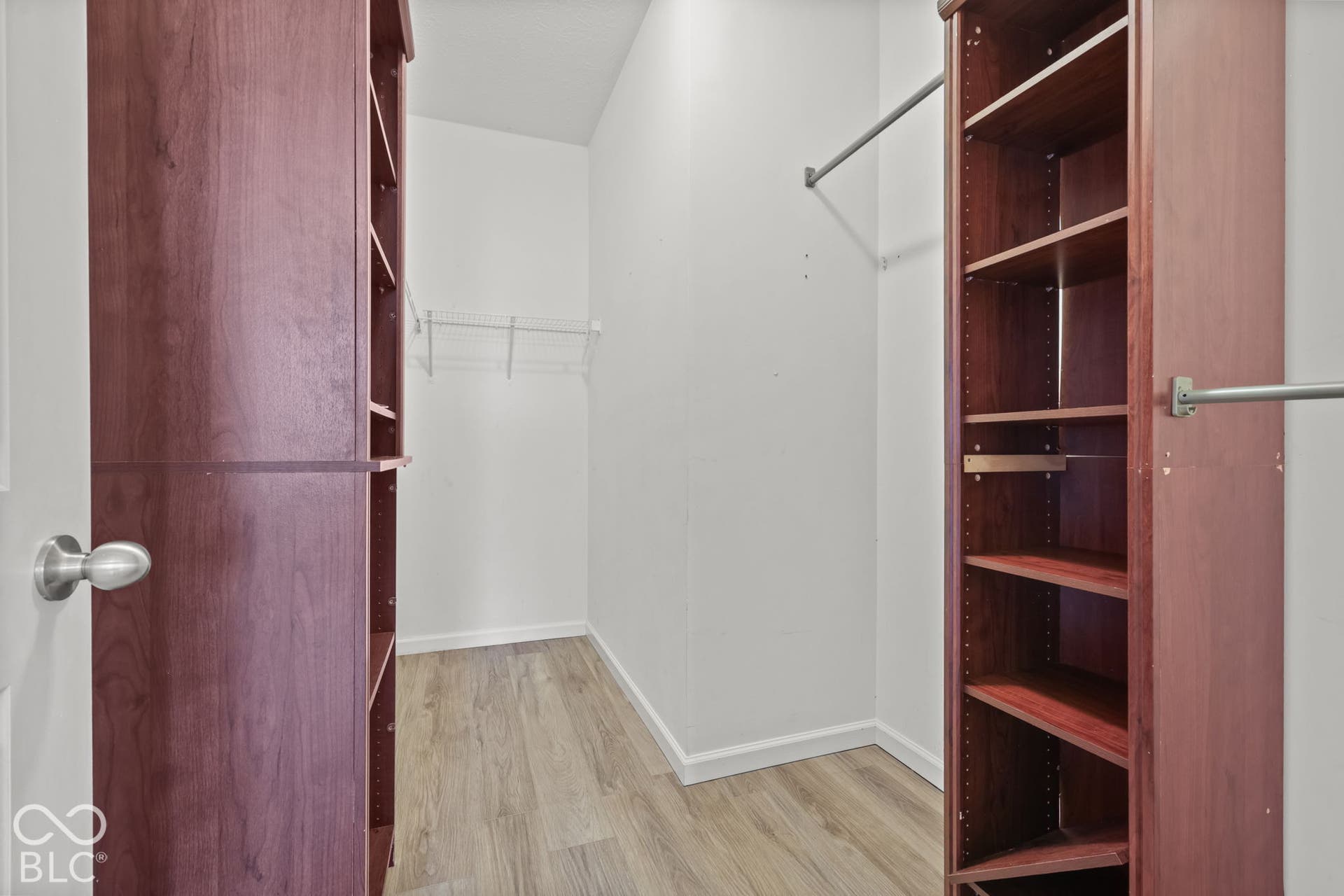 Walk-in storage space featuring wood-finish flooring and floor-to-ceiling built-in shelving units with metal garment rods