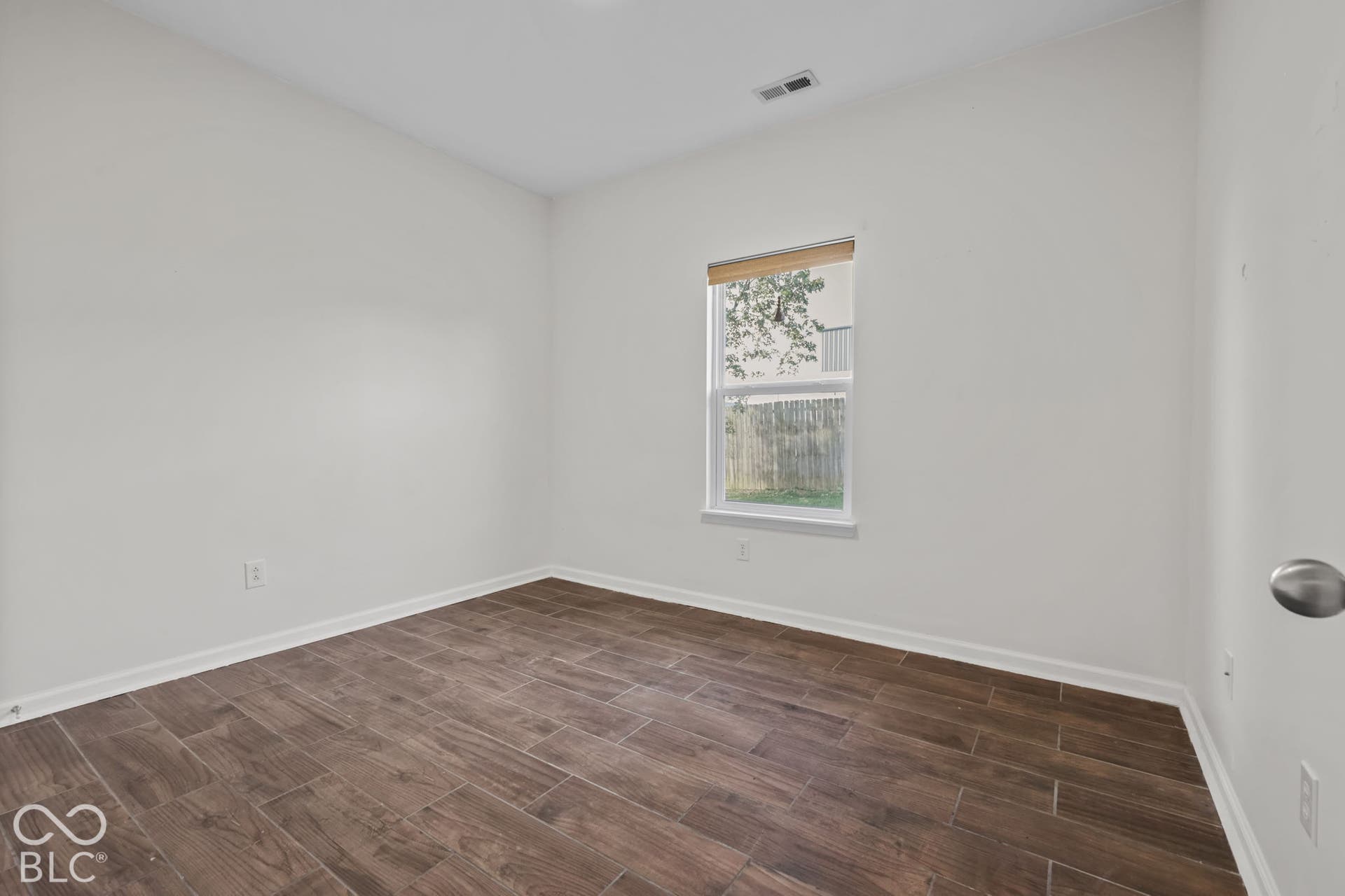 Interior room featuring wood-finish flooring, neutral walls, a single-pane window with a roller shade, and a brushed-nickel door handle