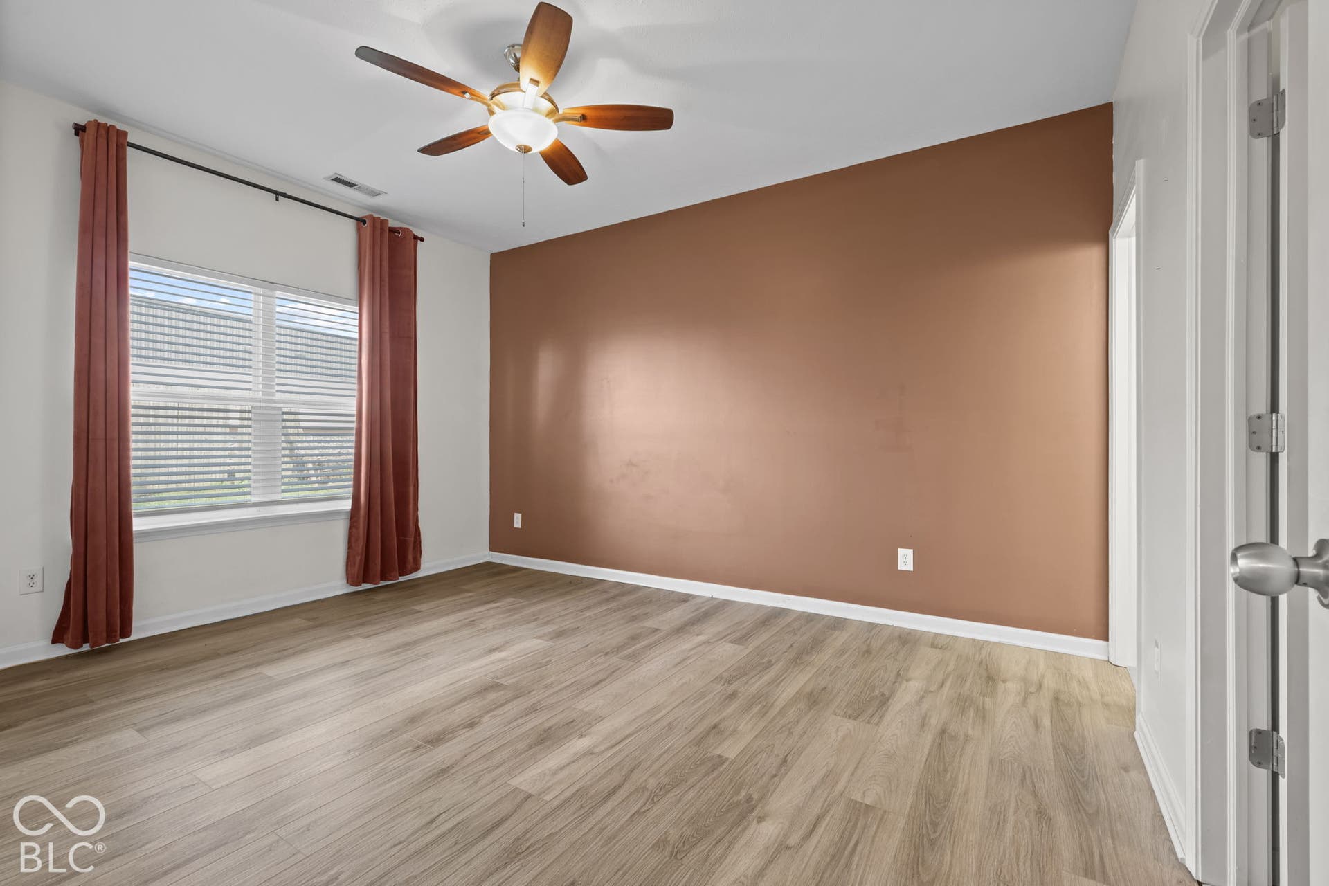 Room featuring wood-finish flooring, a neutral-toned accent wall, a multi-pane window, and a central ceiling fan with integrated lighting