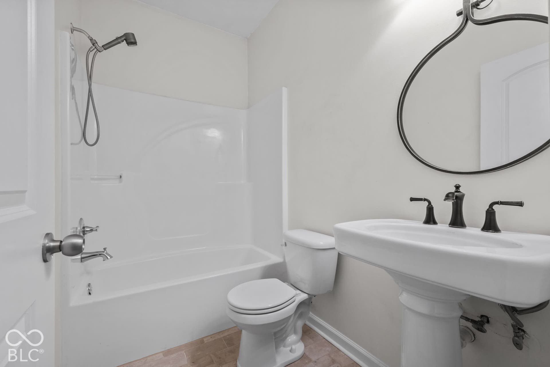 Pedestal sink with dark metal hardware, oval wall-mounted mirror, white bathtub surround, and handheld shower fixture
