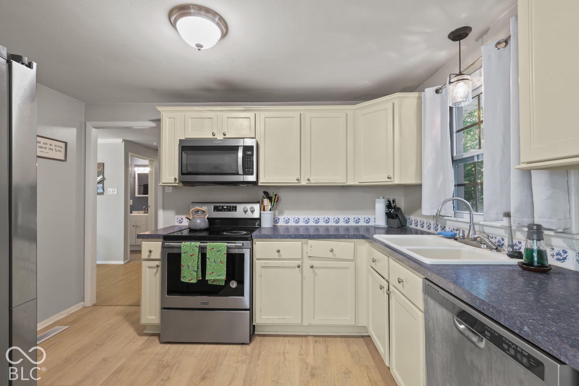 Kitchen featuring stainless steel appliances, shaker-style cabinetry, a dual-basin white sink, patterned tile backsplash, and light-tone wood-finish flooring