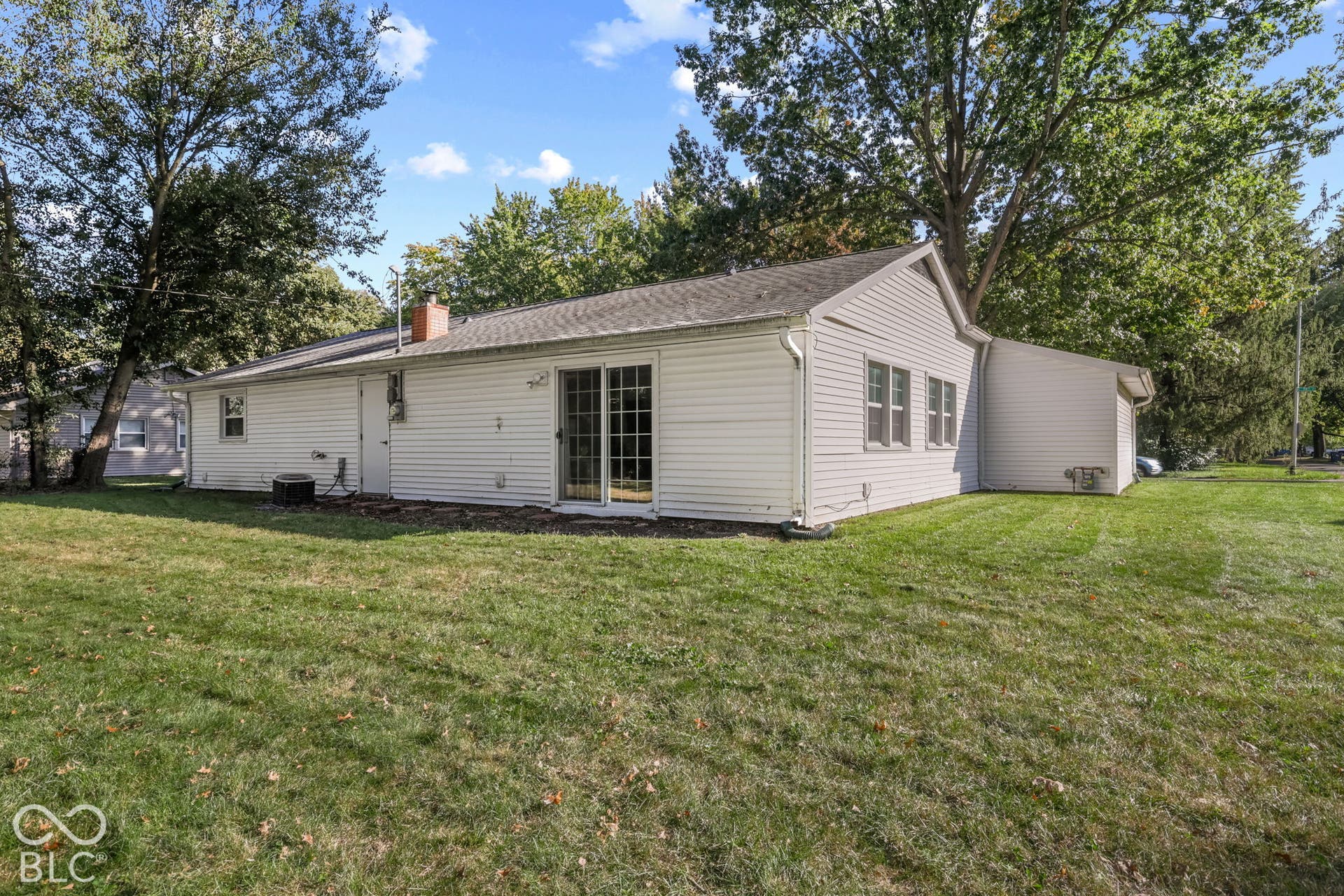 Rear exterior featuring light-colored horizontal siding, sliding glass doors, a brick chimney, and an expansive lawn with mature trees