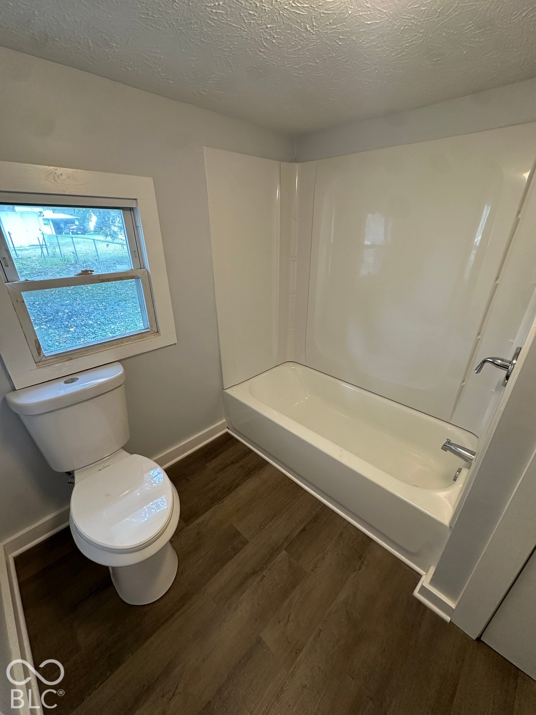 Wood-finish flooring, white bathtub with integrated shower surround, and a toilet with a traditional design