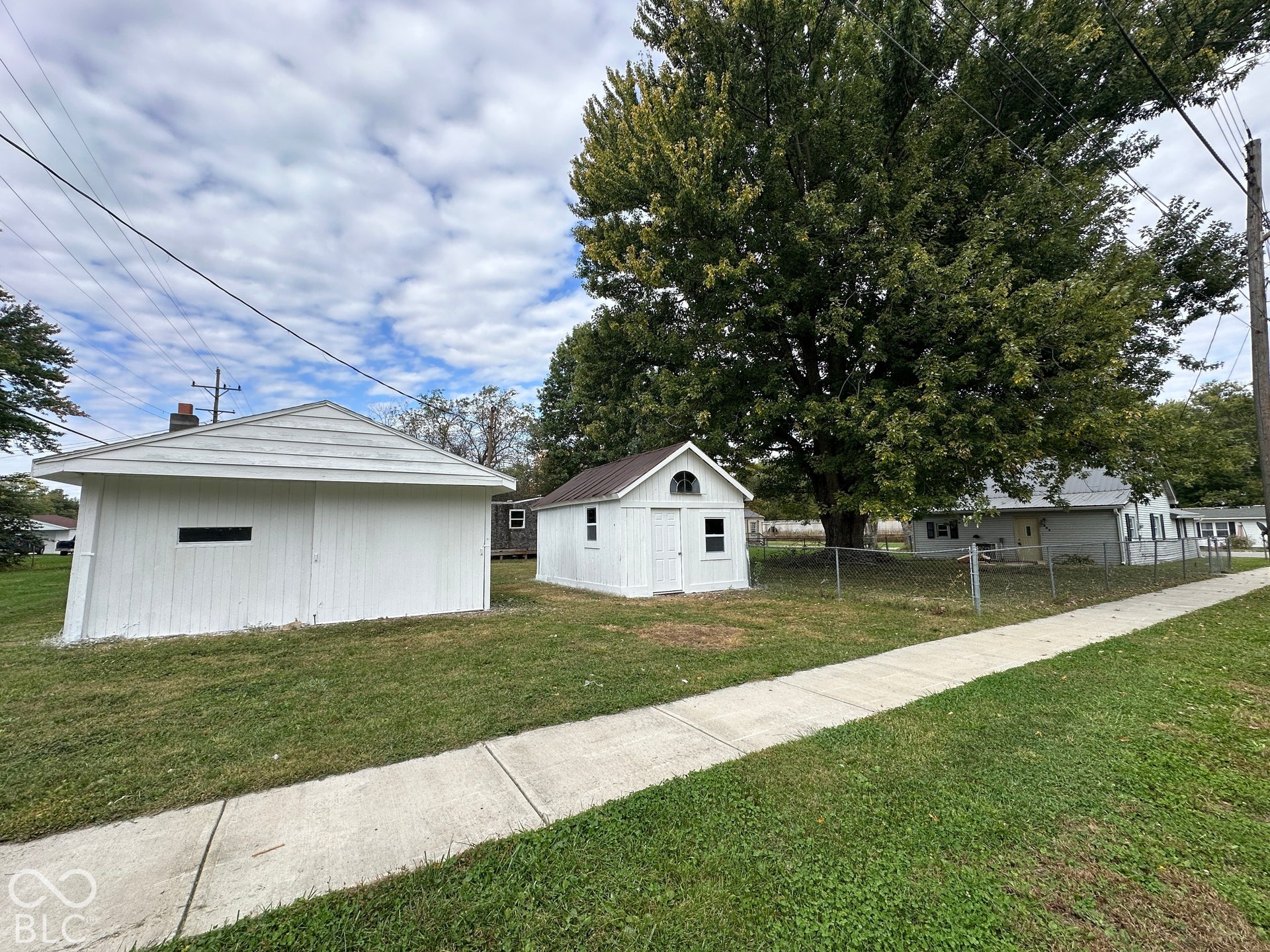 Detached garage with horizontal siding, a separate small shed, established sidewalk, and a chain-link fence