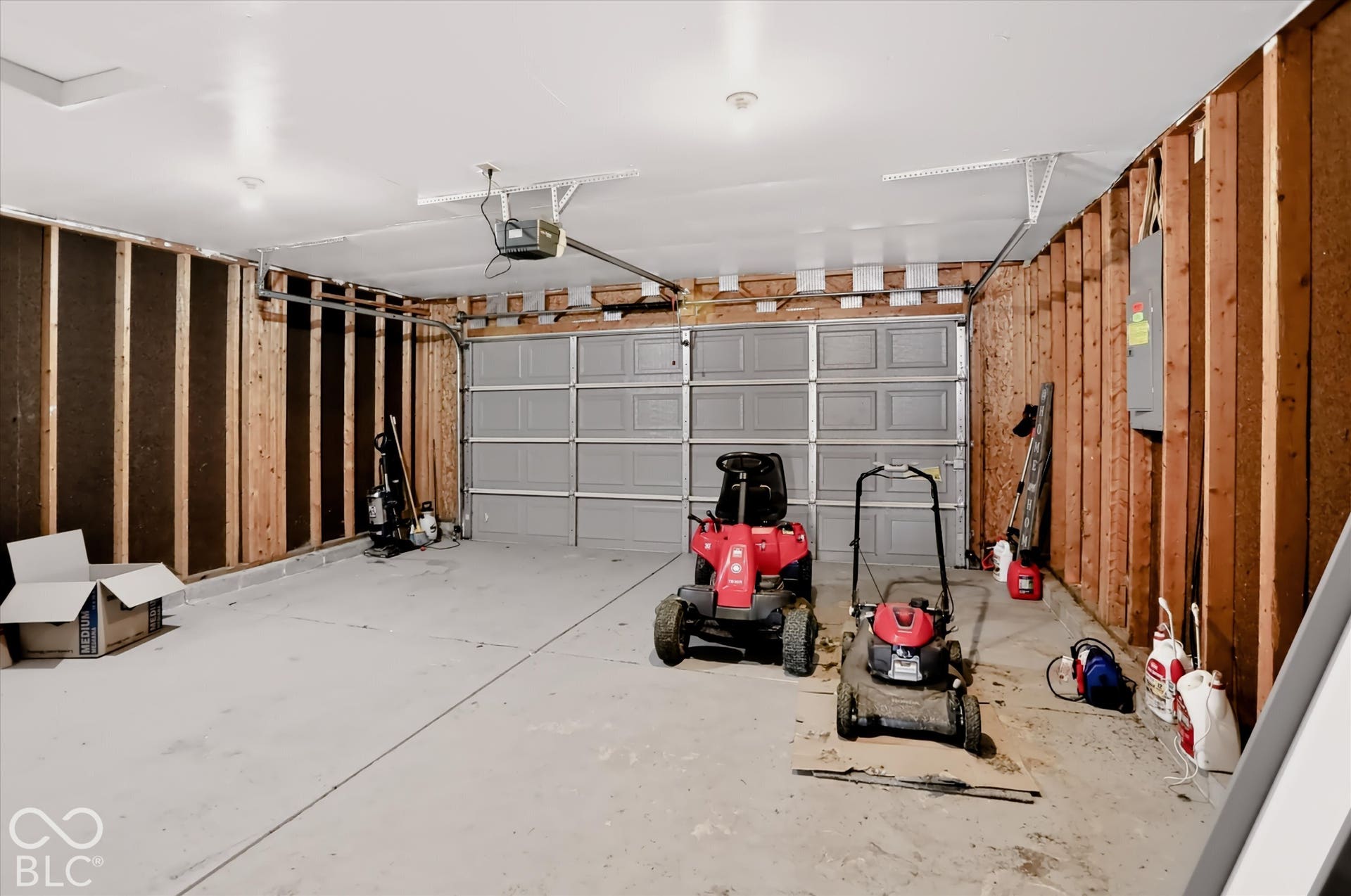 Two-car garage featuring a concrete floor, metal overhead door, electrical panel, and exposed wood-stud wall framing