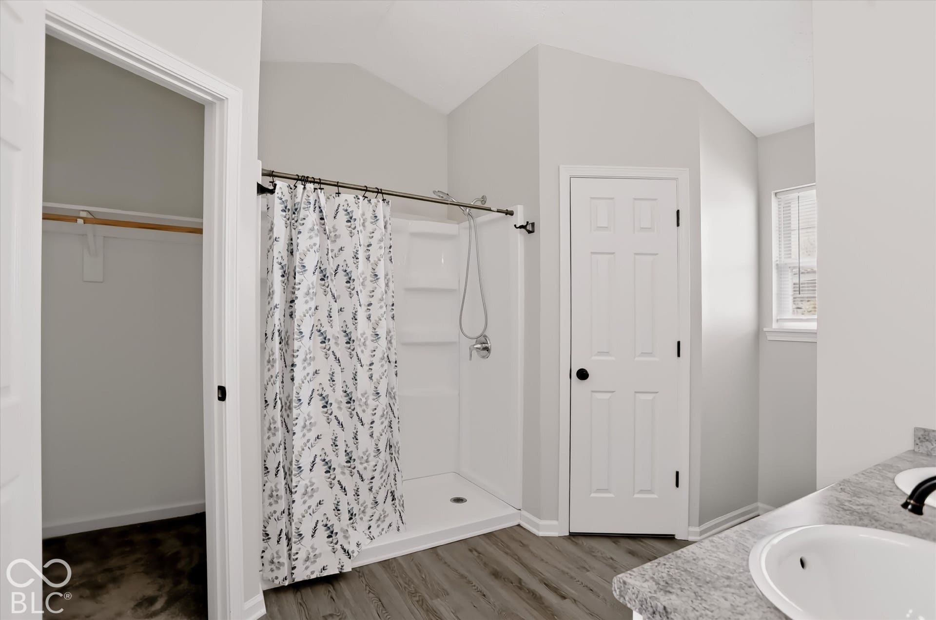 Bathroom featuring a vaulted ceiling, wood-finish flooring, dual-sink vanity with stone-finish countertop, walk-in shower with integrated shelving, and a built-in closet
