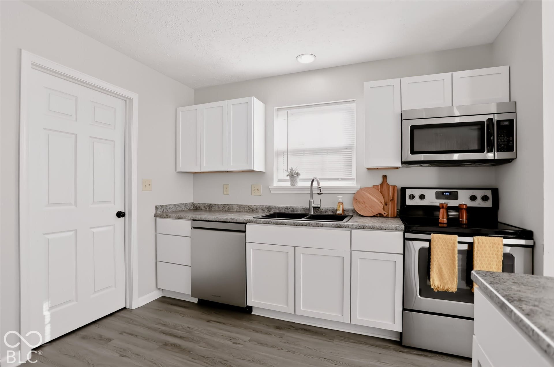 Kitchen featuring white shaker-style cabinetry, stone-pattern laminate countertops, stainless steel appliances, a dual-basin sink, and wood-finish flooring
