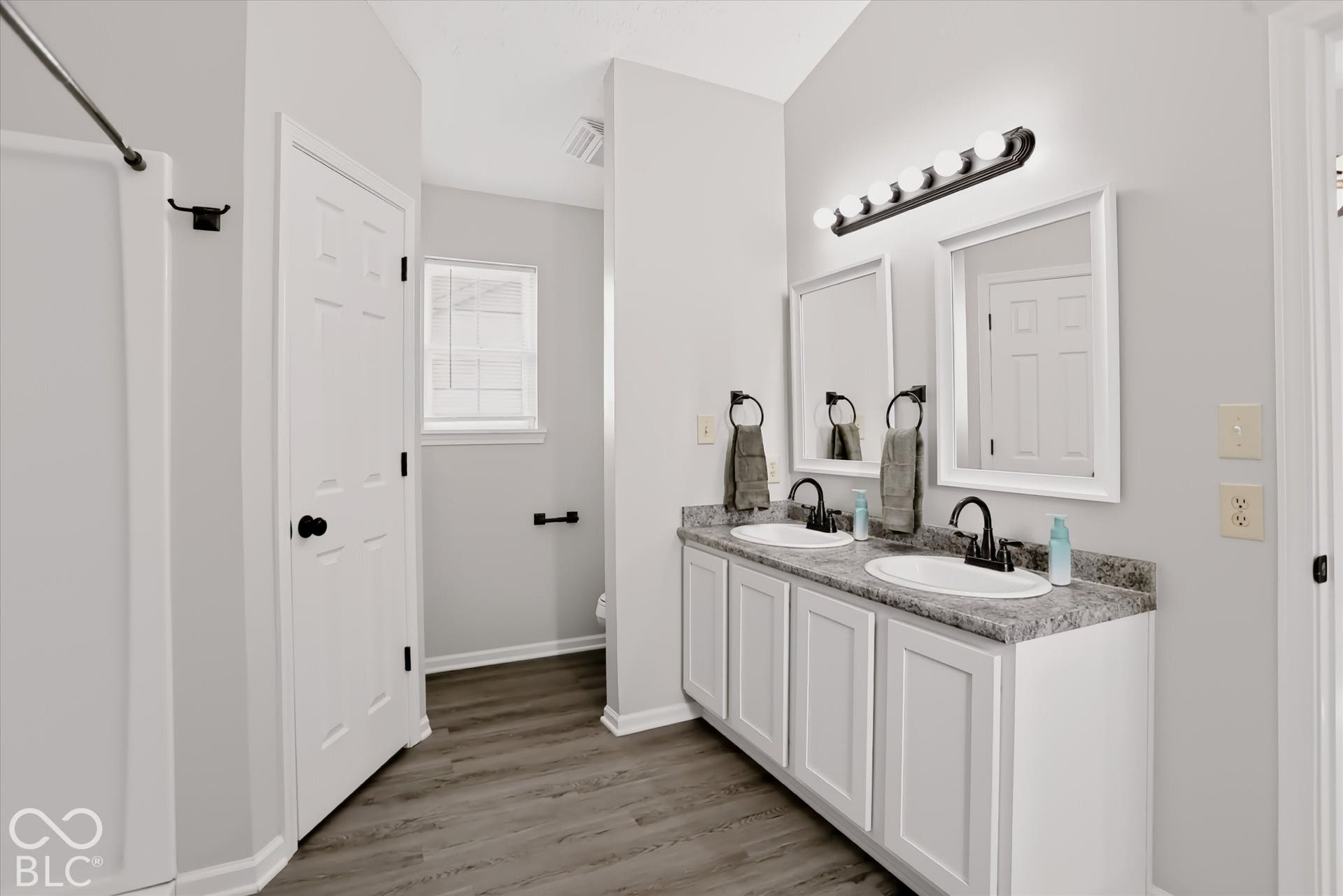 Dual-sink vanity with stone-finish countertop, white shaker-style cabinetry, matte black hardware, and wood-finish flooring