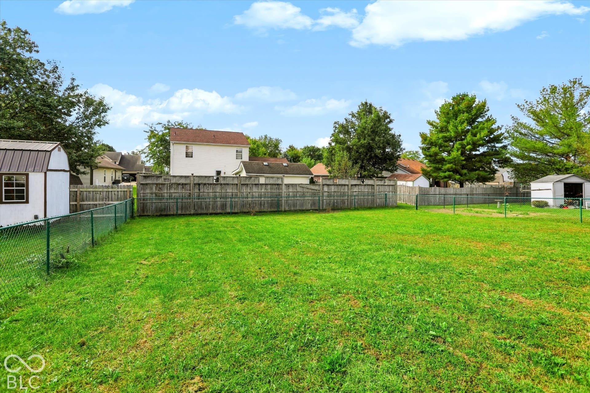 Expansive backyard featuring a fenced perimeter, grass lawn, mature trees, and detached storage structures