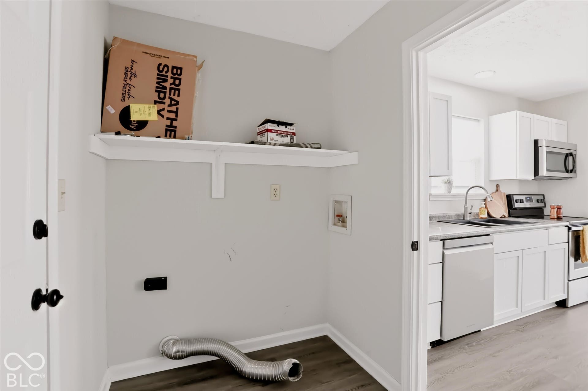 Utility area featuring wood-finish flooring and a wall-mounted shelf, with a doorway providing a sightline into a kitchen equipped with white cabinetry, stainless steel appliances, and quartz countertops