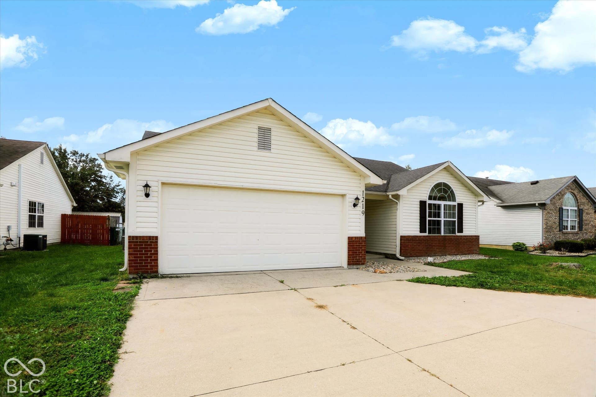 Single-story residence featuring light-colored horizontal siding, brick masonry skirting, an attached two-car garage, and a decorative arched window with grid mullions