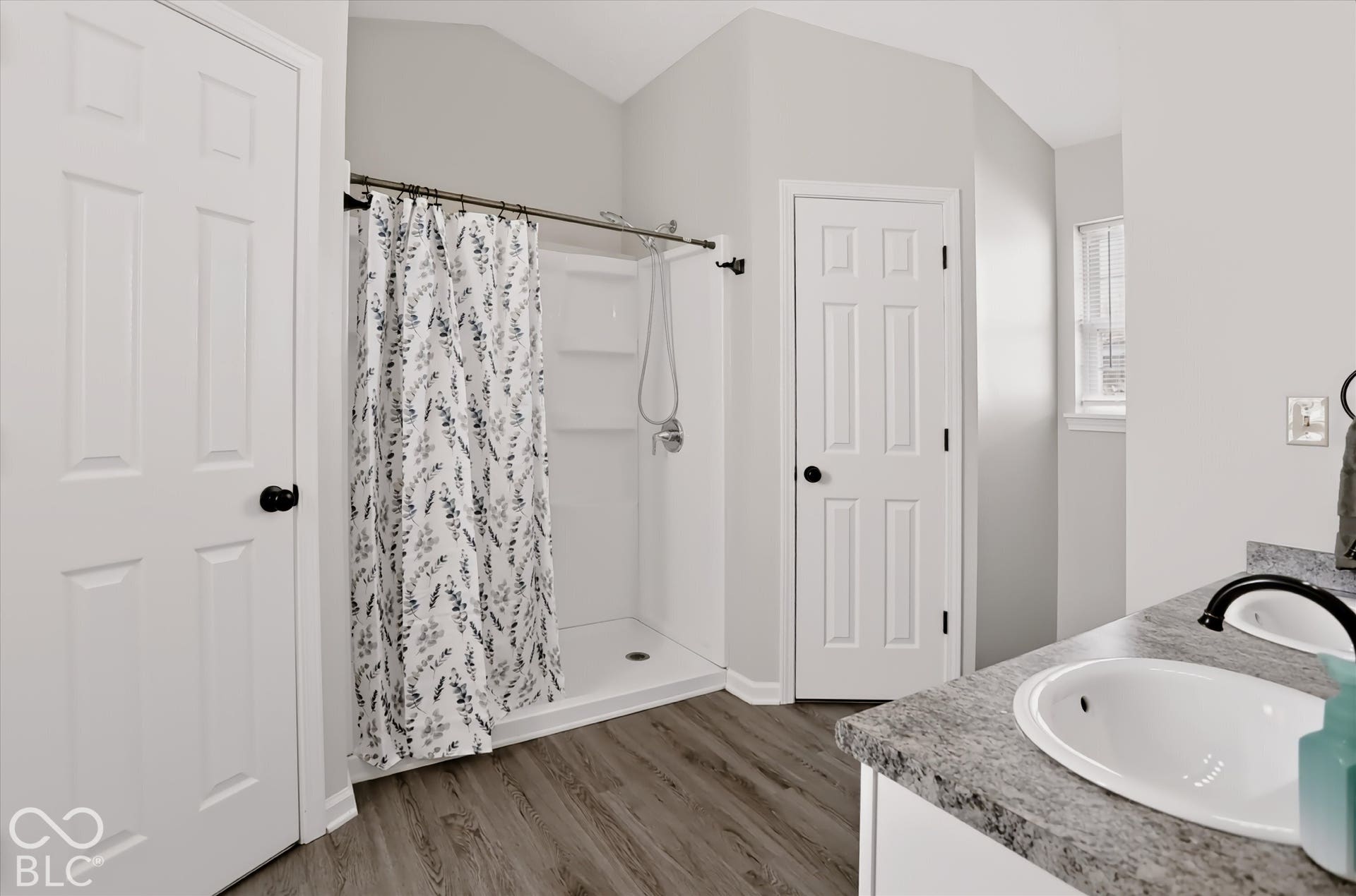 Bathroom featuring a walk-in shower with integrated shelving, dual-sink vanity with stone-finish countertop, matte black fixtures, vaulted ceiling, and wood-finish flooring