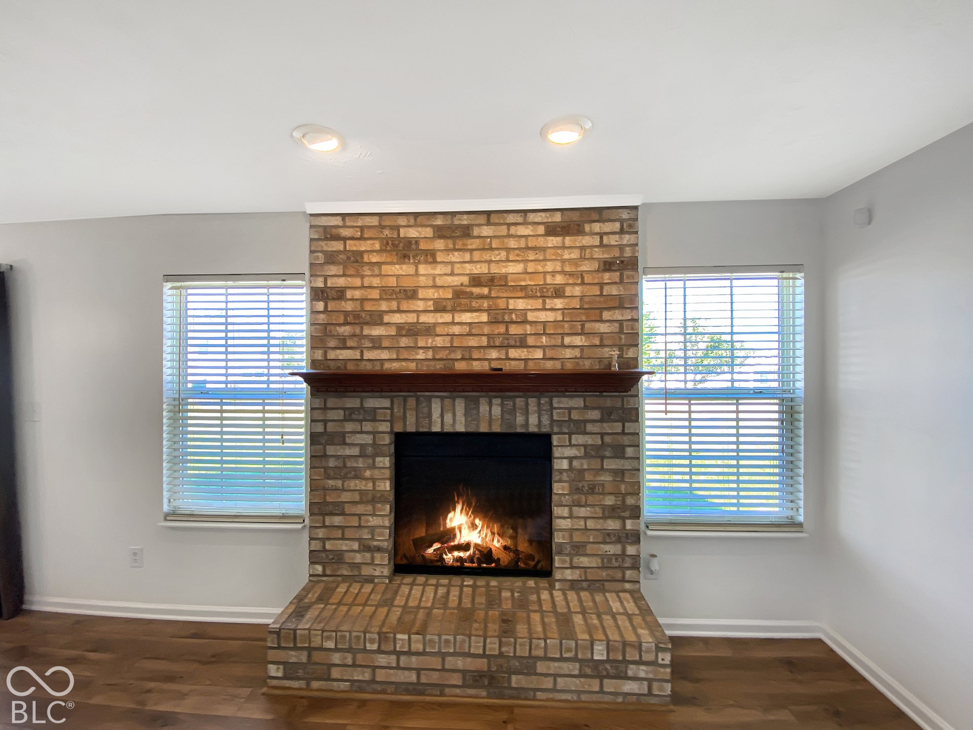4726 Eva Court, Indianapolis, IN 46227 Floor-to-ceiling brick fireplace with a wood mantel and raised hearth, flanked by dual windows with blinds and wood-finish flooring