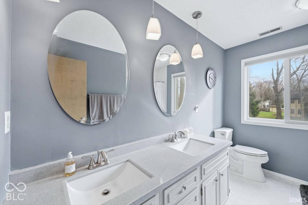 Dual-sink vanity with a solid-surface countertop, twin oval wall-mounted mirrors, dual pendant lighting, and white cabinetry
