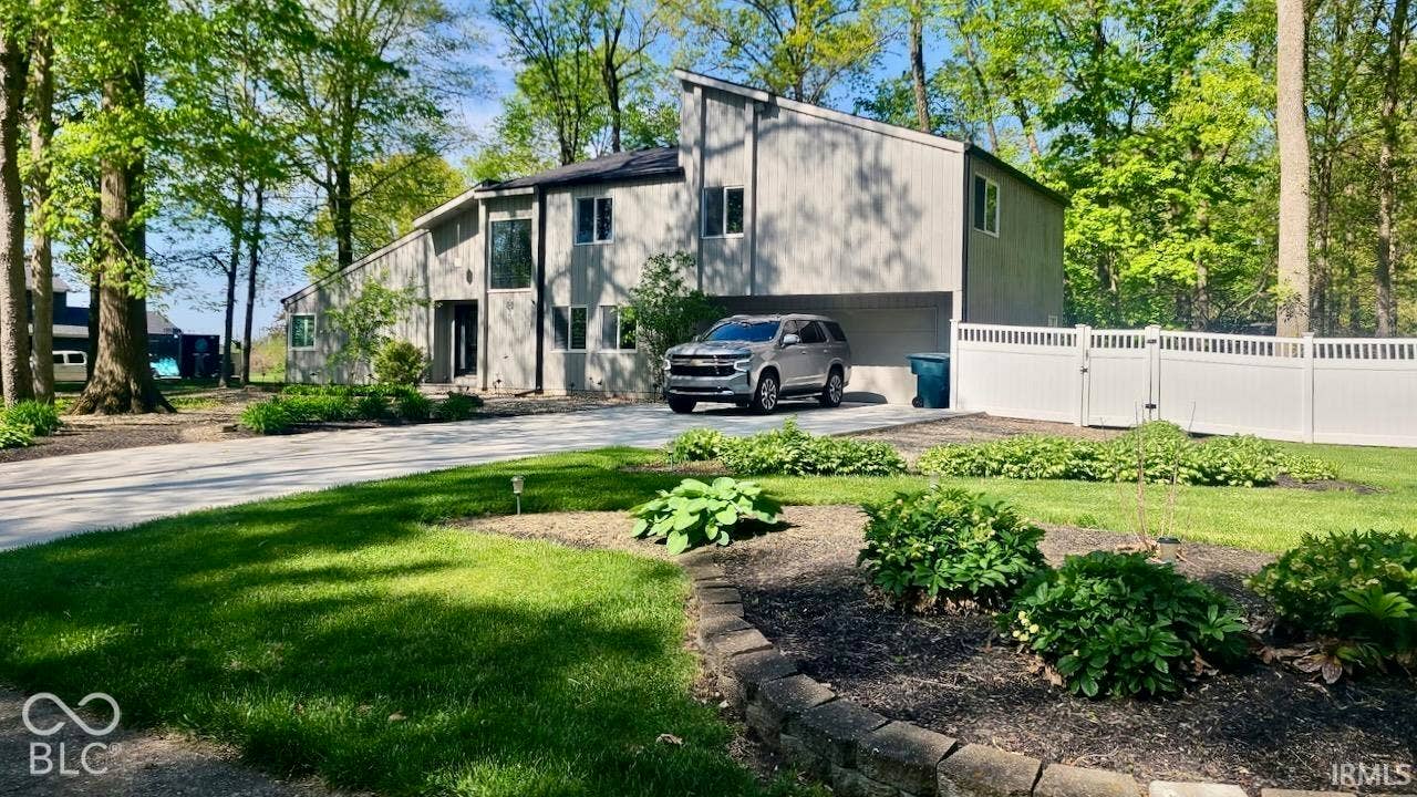 Contemporary exterior featuring vertical siding, a covered parking area, concrete driveway, stone-edged garden beds, and white vinyl privacy fencing
