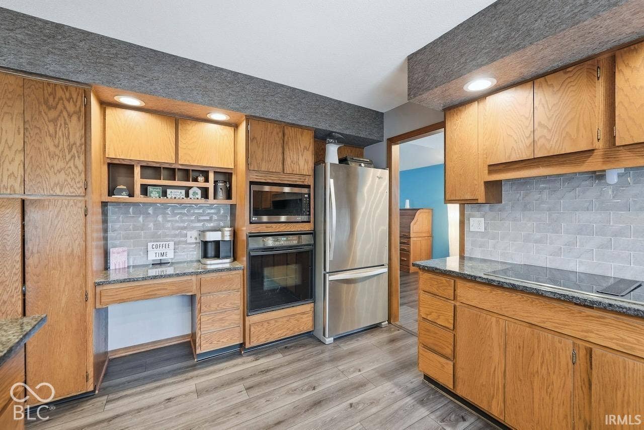 Kitchen featuring wood cabinetry, stone-finish countertops, stainless steel appliances including a built-in oven and microwave, subway tile backsplash, and wood-finish flooring