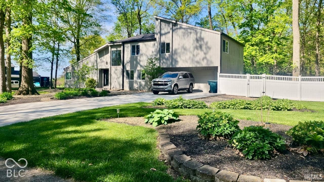 Multi-level residence featuring light gray vertical siding, a paved driveway, an integrated carport, and a white privacy fence, set on a landscaped lot with mature trees